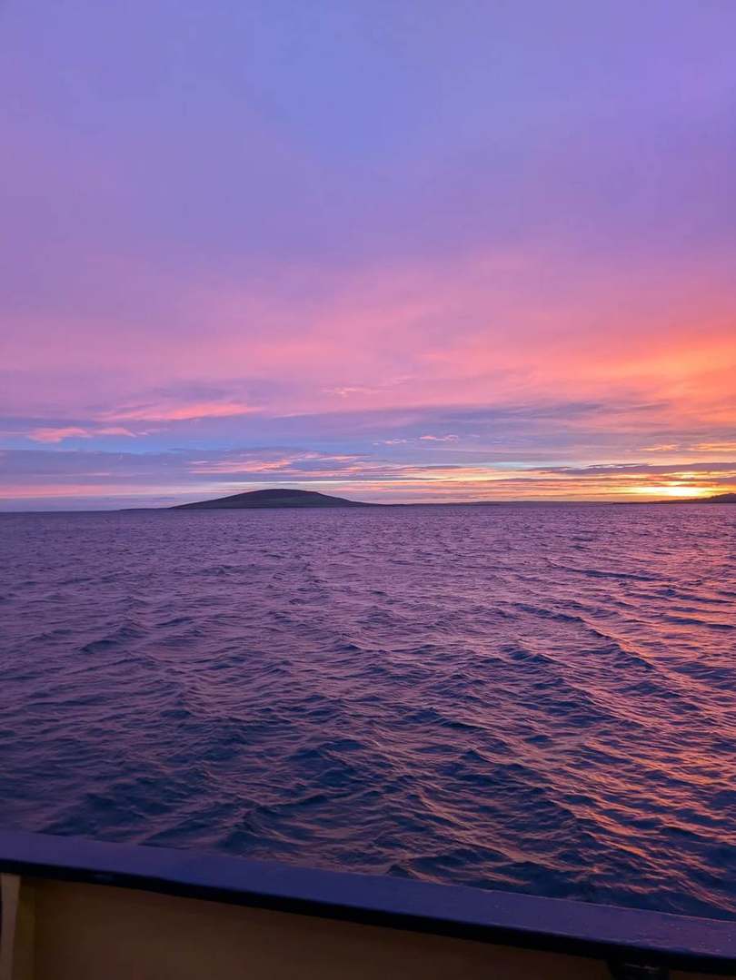 Pink and orange dawn sky over the sea and a low island, seen from the ferry