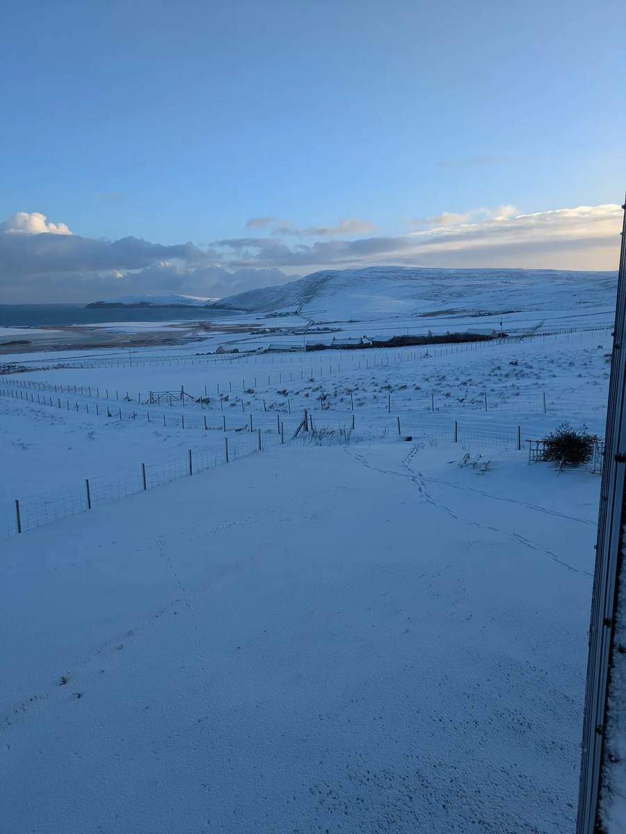 Footprints through snowy fields on Rousay, with fences, buildings, a coastal hill and the sea beyond