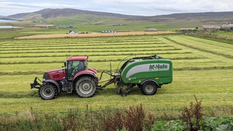 A red tractor and green baler in a field of cut grass on Rousay, with farm buildings and hills beyond