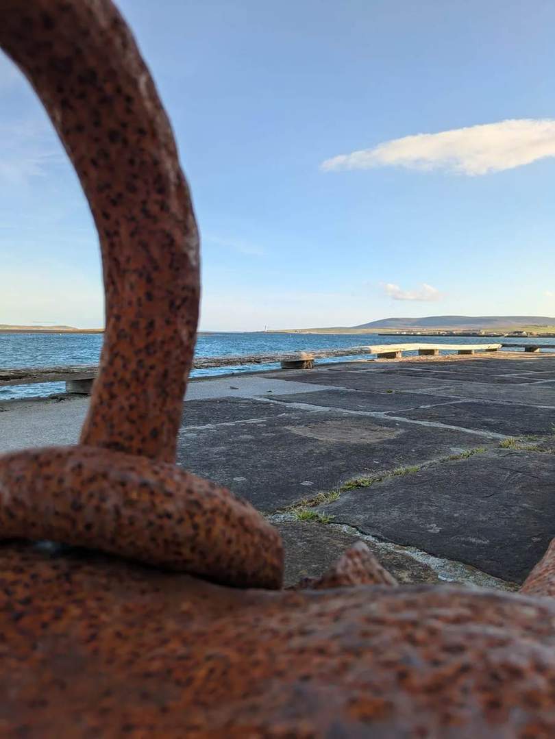 Sea and low hills seen from a stone quay in Stromness, framed by rusty ironwork