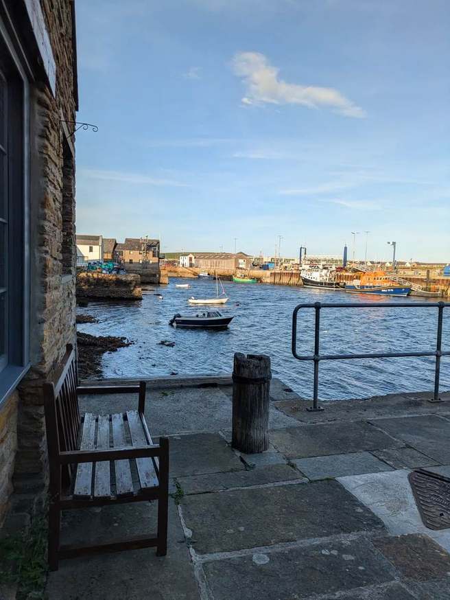 Stone waterfront building and bench beside boats in Stromness harbour, with an orange lifeboat beyond
