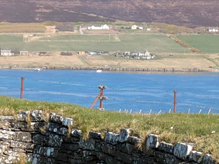 View from Wyre across blue water to fields and hillside houses, with a stone wall in the foreground